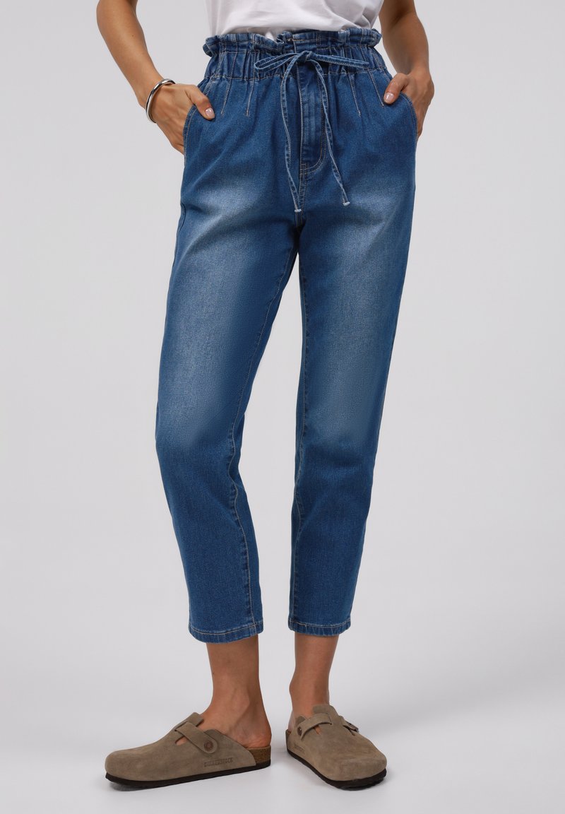 Blå denimbyxor med en samlad, dragkedja midjeresår och sidofickor; passar löst med uppvikta anklar; kombineras med bruna slip-on skor.