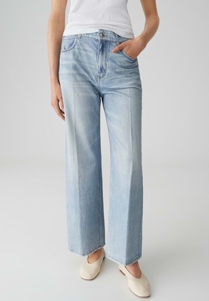 Vrouw die lichtblauwe jeans met wijde pijpen, een witte mouwloze top en beige instapschoenen draagt, staand met één hand in de zak.