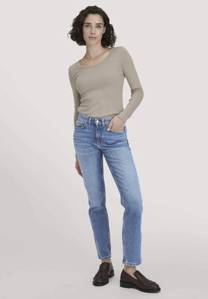 Vrouw met kort, donker krullend haar, gekleed in een beige lange mouw top, lichtblauwe jeans en bruine loafers, staande met één hand in de zak.