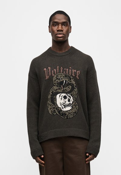 Νέος άνδρας που φοράει μια σκοτεινή πλεκτή μπλούζα με το κείμενο «Voltaire» και ένα μεγάλο κεντημένο φίδι που είναι τυλιγμένο γύρω από ένα κρανίο στο μπροστινό μέρος.