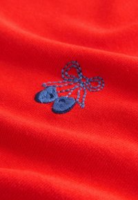 Tela roja con una textura suave que presenta un lazo bordado en azul y pequeños detalles de colgante, creando un elemento de diseño distintivo.