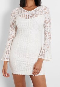 Robe mini en dentelle blanche avec de longues manches évasées, présentant un motif texturé et des panneaux transparents. Doublure lisse sous la superposition en dentelle.