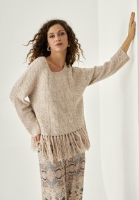 Suéter de punto beige con patrón de trenzas y dobladillo con flecos, hecho de hilo suave. Llevado sobre pantalones estampados con diseños florales y en espiral.