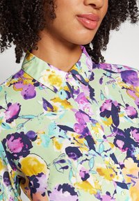 Camisa de botão floral com um fundo verde claro e flores roxas, cor-de-rosa e amarelas; com colarinho clássico; e botões verdes claros.