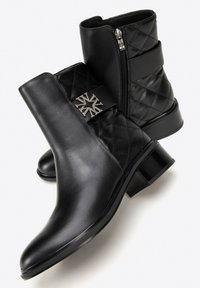 Botas negras hasta el tobillo con un frente de cuero liso, parte trasera acolchada, cremallera lateral y un adorno metálico en la correa. Diseño de tacón grueso.