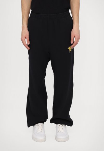 Pantalon de survêtement noir en tissu doux, présentant une coupe décontractée, une taille élastique et des accents de baskets blanches. Détail du logo doré sur le côté.
