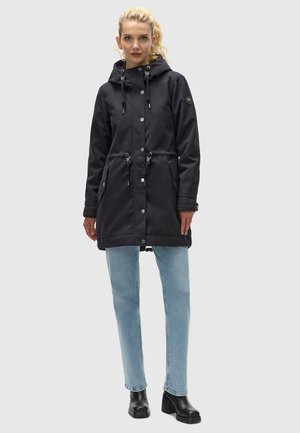 Parka noire à manches longues avec taille à cordon et capuche, dotée de boutons-pression et de deux poches avant, associée à un jean bleu clair et des bottes noires.