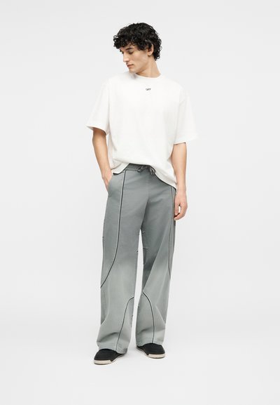 Νεαρός άνδρας που φοράει λευκό φαρδύ T-shirt και άνετο γκρι παντελόνι με μαύρα ραφές, στέκεται με τα χέρια στις τσέπες σε απλό φόντο.