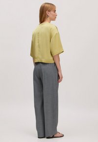 Satin-gelbes Cropped-Top mit weiten kurzen Ärmeln, kombiniert mit lockeren grauen Karohosen. Das Model steht mit dem Rücken zur Kamera.