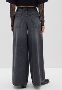 Jeans de pierna ancha en denim gris oscuro con cintura elástica, que presentan bolsillos traseros y un dobladillo deshilachado. La tela tiene un acabado texturizado.