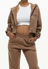 Hnědá krátká mikina s kapucí a odpovídající tepláky z měkké látky. Mikina má přední zip a logo, kalhoty mají elastický pas a kapsy.