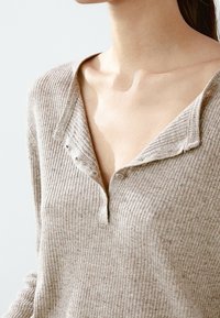 Beige ribbed sweater with a V-neck and four-button placket. The fabric is textured, featuring subtle variations in color and thickness.