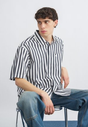 Νεαρός άνδρας με σγουρά μαλλιά που φοράει κοντομάνικο πουκάμισο με κάθετες μαύρες και λευκές ρίγες και μπλε τζιν, κάθεται σε μεταλλική καρέκλα με φόντο λευκό.