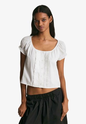 Bílá krátká halenka s hranatým výstřihem, balónovými rukávy a knoflíkovým zapínáním vpředu s volánkovými detaily uprostřed a na manžetách.