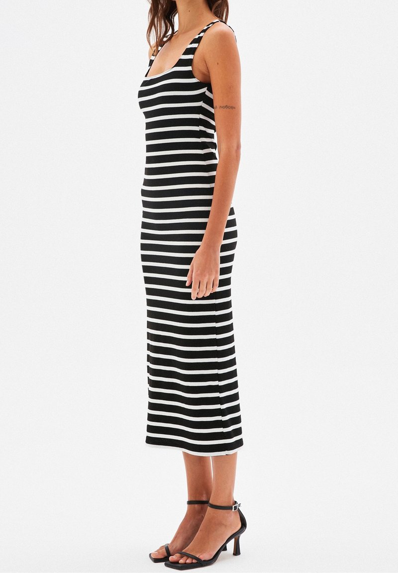 Femme portant une robe midi sans manches à rayures horizontales noires et blanches avec des sandales noires à talons hauts et bride à la cheville, debout de profil.