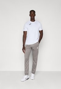 Witte sportieve T-shirt met korte mouwen en zwart logo, gecombineerd met lichtgrijze joggingsbroek met witte branding. Model draagt witte sportschoenen.