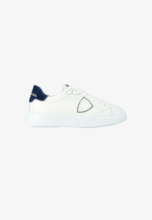Baskets blanches basses avec un talon arrière bleu marine et une semelle en caoutchouc blanc arrondi, présentant un système de laçage et un logo discret cousu sur le côté.