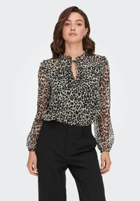 Blouse en imprimé léopard avec des manches longues en tulle, un décolleté en forme de fenêtre avec un détail à nouer, et une coupe légèrement ample. Portée avec un pantalon noir.