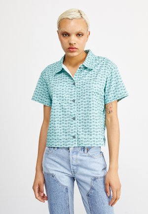 Kurzärmliges Hemd mit Knöpfen in hellem Türkis mit geometrischem Muster. Verfügt über einen Kragen und ein verkürztes Design. Kombiniert mit hellblauen Jeans.