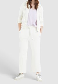 Kvinde iført off-white zip-up hoodie, matchende brede sweatpants, lyslilla top og hvide sneakers, stående med hænderne i lommerne.