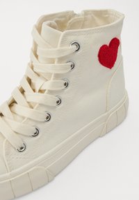 Högre canvas sneakers i off-white, med ett texturerat rött broderat hjärta på sidan, beige snören och en gummisula.