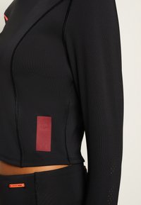 Schwarzes Langarmoberteil mit tailliertem Design, das rote Akzente und ein strukturiertes Logo-Patch an der Seite aufweist. Mesh-Details an den Ärmeln verbessern die Atmungsaktivität.