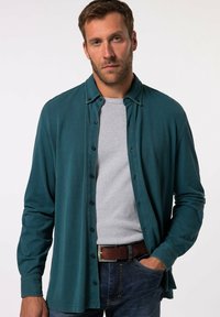 Camicia verde acqua con maniche lunghe sopra una maglietta grigia, indossata con jeans blu, dotata di una cintura marrone e un fit rilassato.