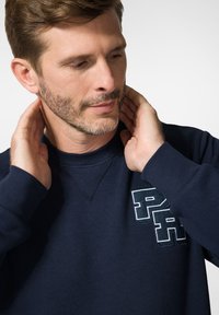 Granatowy sweatshirt z okrągłym dekoltem i teksturowanym materiałem; z wyraźnym haftowanym logo na lewej klatce piersiowej.