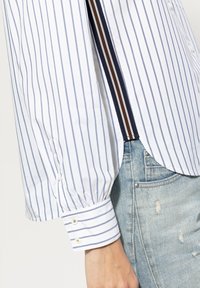 Striped shirt in white and navy with a relaxed fit, featuring a buttoned cuff and side split detail, paired with distressed light blue jeans.