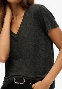 Dunkelgraues T-Shirt mit kurzen Ärmeln und V-Ausschnitt sowie subtilen goldenen Sprenkeln; kombiniert mit schwarzen Hosen und einem schwarzen Gürtel mit goldener Schnalle.