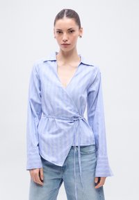 Ανοιχτό μπλε πουκάμισο τύπου wrap με κάθετες λευκές ρίγες. Διαθέτει βαθύ V-λαιμόκοψη, μακριά μανίκια με φαρδιές μανσέτες και λεπτομέρεια δεσίματος στη μέση.