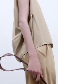 Top beige senza maniche realizzato in tessuto leggero, con una vestibilità rilassata e spacchi laterali. Cintura in pelle marrone tenuta in mano, abbinata a pantaloni beige.