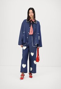 Denim jacket with contrast white stitching, matching denim pants featuring white heart cutouts, and a red handbag. Striped shirt underneath.