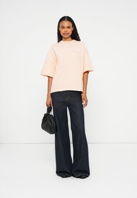 T-shirt oversized rosa claro com mangas curtas, combinado com jeans de perna larga azul-escuro. O modelo segura uma bolsa preta com pregas.