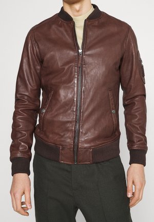 Mężczyzna noszący brązową skórzaną kurtkę bomber z przednim zamkiem błyskawicznym, ściągaczami na rękawach i u dołu oraz bocznymi kieszeniami, na beżowej koszuli i ciemnych spodniach.