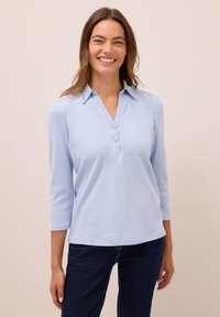 Uśmiechnięta kobieta w jasnoniebieskiej koszuli z kołnierzykiem zapinanej na 3 guziki z rękawami 3/4 i ciemnoniebieskich dżinsach, stojąca na neutralnym tle.