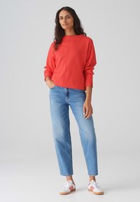 Sweatshirt rouge à manches longues, col rond, avec des épaules tombantes, associé à un jean bleu clair à coupe ample et des baskets blanches avec des rayures rouges.