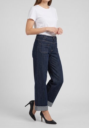 Donna che indossa una t-shirt bianca a maniche corte, jeans blu scuro risvoltati e scarpe nere con tacchi alti, in piedi davanti a uno sfondo semplice.