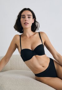 A woman with short, curly hair poses on a light textured surface, wearing a black bra and matching panties, conveying fashion and comfort.