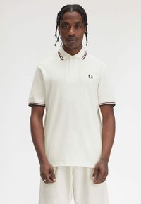 Polo bianco con maniche a strisce nere e bordeaux, dotato di un piccolo logo ricamato sul petto sinistro. Tessuto morbido, vestibilità classica.