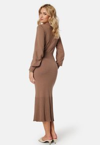 Robe midi tricotée marron avec une texture côtelée, silhouette ajustée et longues manches bouffantes. Elle présente un ourlet évasé et un col montant.