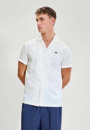 Joven con cabello corto, vestido con una camisa blanca de manga corta y botones, y gafas de sol en la cabeza, mirando hacia la izquierda sobre un fondo liso.