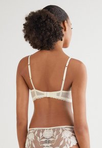 Crème kant bh met verstelbare bandjes, een striksluiting en geschulpte randen; heeft semi-doorzichtig materiaal en een delicate, gestippelde patroon.