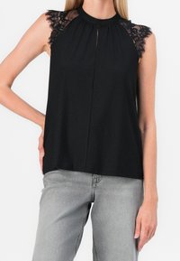 Top nero senza maniche con dettagli in pizzo sulle spalle, apertura a chiave sul davanti e vestibilità morbida. Indossato con jeans grigio chiaro.