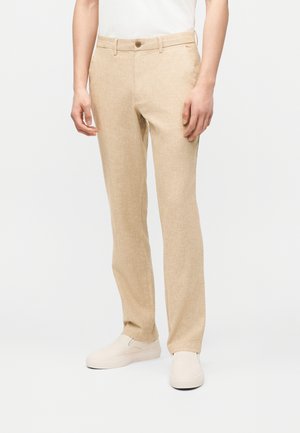 Beige Leinenhose mit Knopf- und Reißverschluss, getragen mit weißen Slippern und einem weißen Hemd an einem stehenden männlichen Modell.