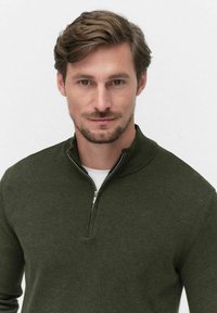 Tummanvihreä pullover- neule, jossa on korkea kaulus ja puolivetoketju edessä. Valmistettu tekstuurillisesti neulottua materiaalia, käytetään valkoisen paidan päällä.