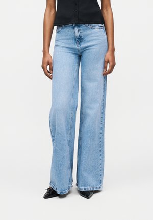 Άτομο που φοράει ανοιχτό μπλε τζιν παντελόνι με φαρδιά πόδια, μαύρα μυτερά παπούτσια και αμάνικο μαύρο πουκάμισο με κουμπιά, στέκεται μπροστά σε απλό φόντο.