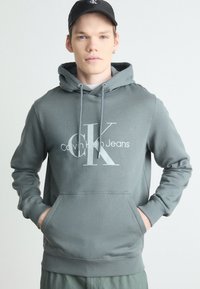 Siva kapuca s kapuco, na kateri je velik beli logo "CK" in napis "Calvin Klein Jeans". Mehak tekstil, kengurujšča žep, ribani manšeti.