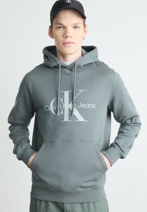 Sudadera gris con capucha, que presenta un gran logo blanco de "CK" y el texto "Calvin Klein Jeans". Tejido suave, bolsillo canguro, puños de canalé.