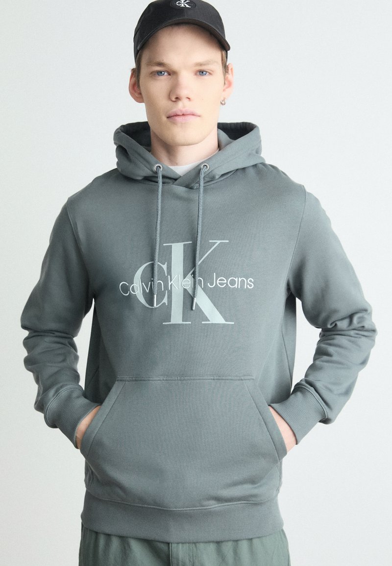 Siva kapuca s kapuco, na kateri je velik beli logo "CK" in napis "Calvin Klein Jeans". Mehak tekstil, kengurujšča žep, ribani manšeti.